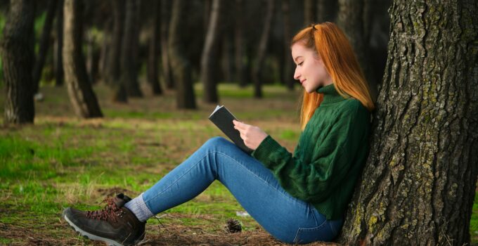 Redhead young woman reading a book sitting under tree in forest.